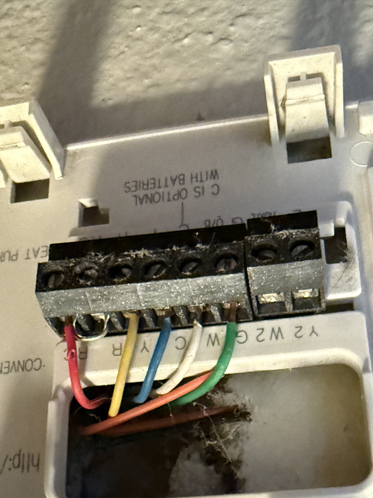 The old Honeywell thermostat back plate with wires still attached. Terminals are labeled Rc, R, Y, C, W and G down the strip, with a red wire on Rc, yellow on Y, blue on C, white on W and green on G. Printed text on the plate reads C IS OPTIONAL WITH BATTERIES.